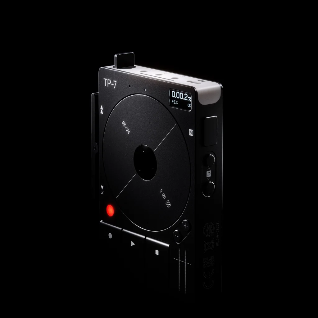 A black Teenage Engineering TP–7 field recorder with a large circular motorized reel, compact controls, and a small display.